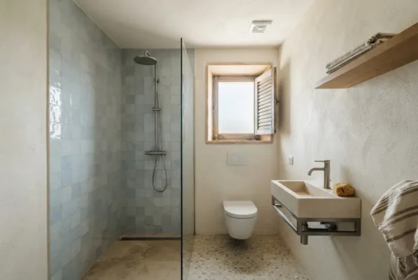 Ein Bad hat eine Walk-in-Dusche mit Glaswand, verkleidet mit hellblauen Fliesen, ein Wand-Waschbecken, ein Wand-WC und einen Terrazzoboden.