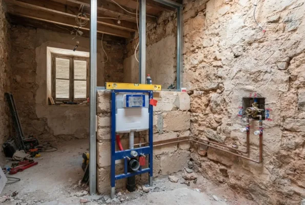 Ein Bad in der Renovierung mit einem Unterputz-Spülkastengestell vor freigelegtem Stein, mit sichtbaren Kupferrohren an der Wand.