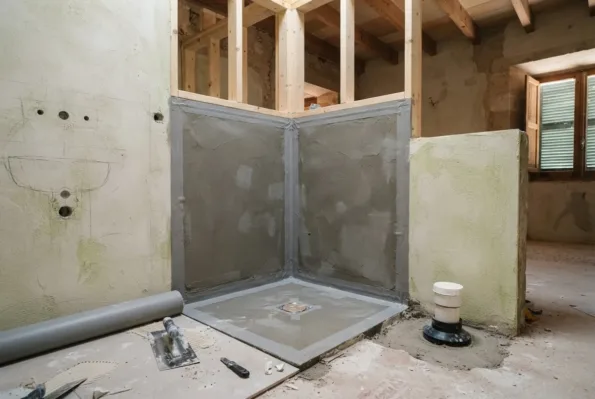 A bathroom mid renovation shows grey waterproofing membrane on new shower walls, a floor drain, a roller, and a trowel.