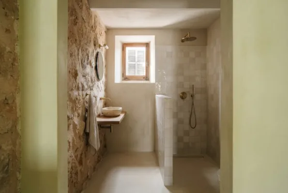 A narrow bathroom set within a room has a stone wall basin and a half height tiled partition before a shower recess.