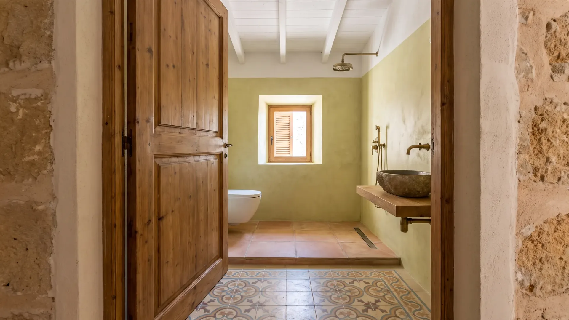An open wood door reveals a pale green bathroom with a terracotta floor, patterned tiles, a wall-hung toilet and stone basin.