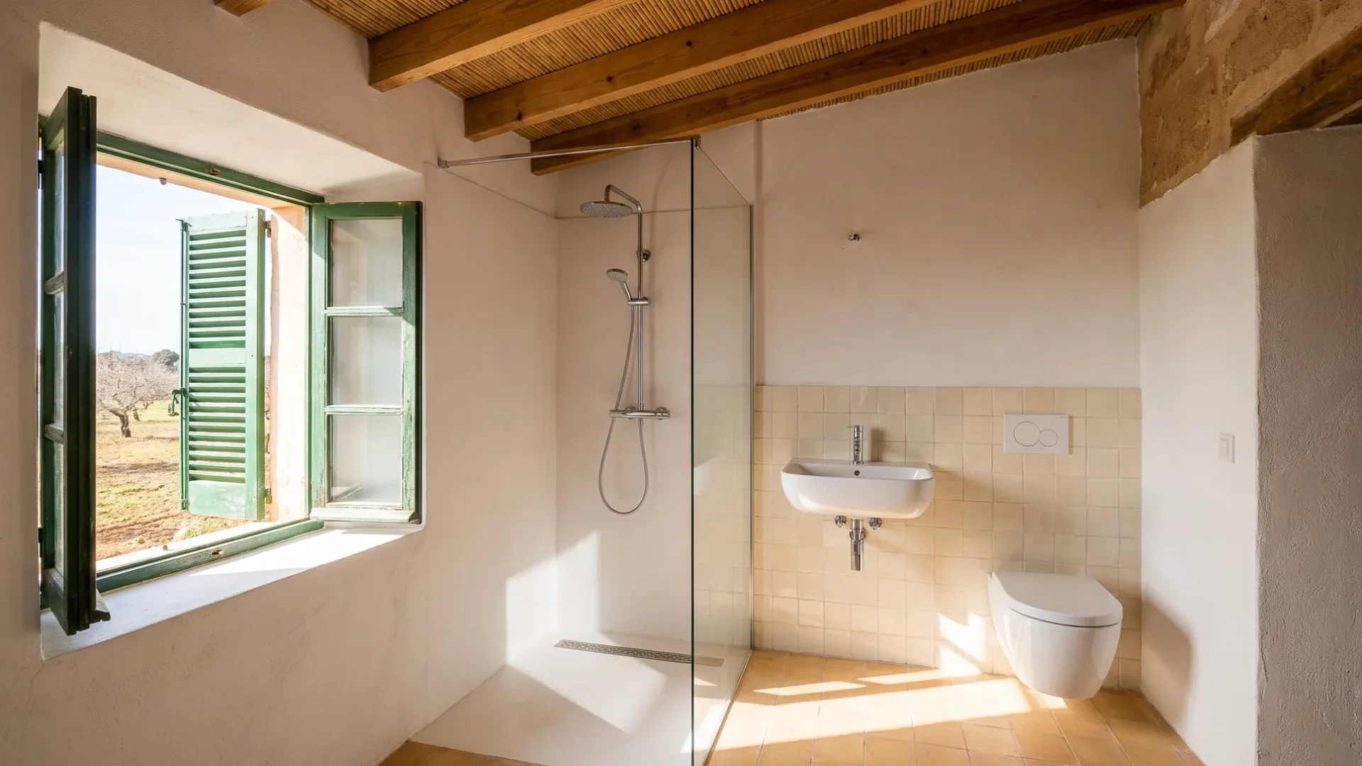 A glass shower stands beside a tiled half-wall with a wall-hung basin and toilet, lit by an open green-shuttered window.
