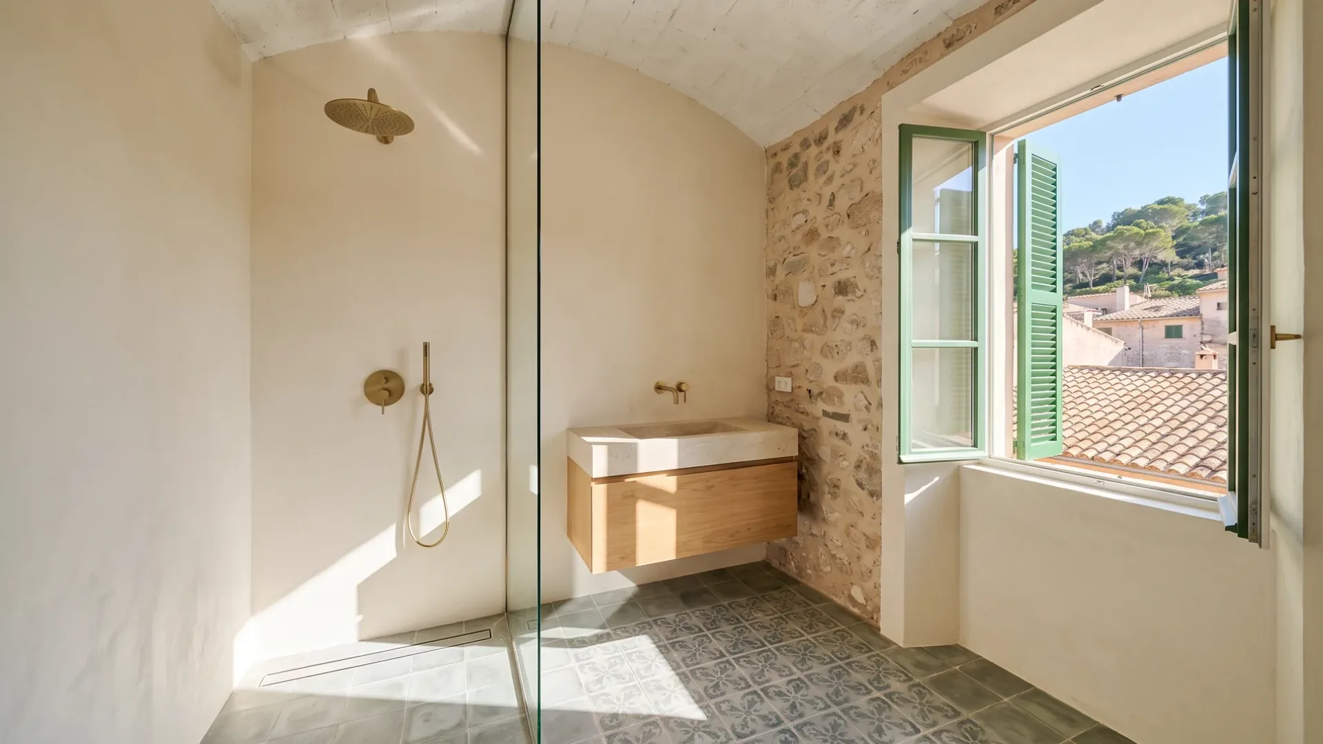 Eine gewölbte Dusche und ein Eichenwaschtisch mit Steinwaschbecken stehen neben einem Fenster mit grünen Fensterläden über Terrakottadächern.