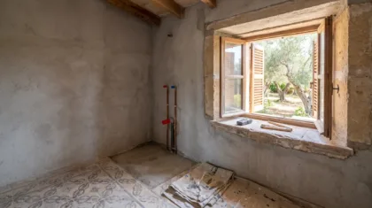 A bathroom stripped for renovation, old patterned floor tiles lifting, capped pipes on bare render, a window onto olives.