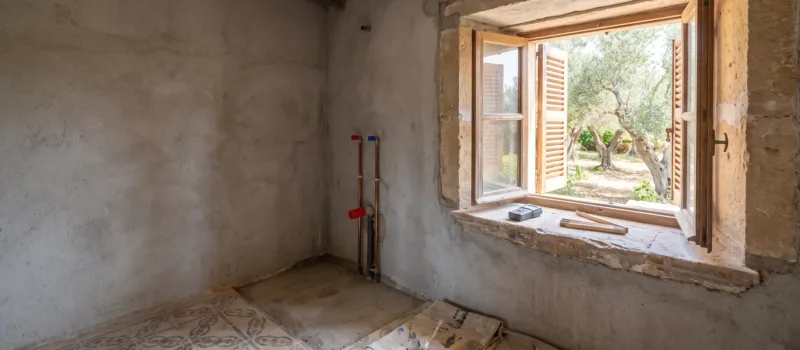 A bathroom stripped for renovation, old patterned floor tiles lifting, capped pipes on bare render, a window onto olives.