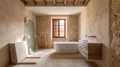 A bathroom mid renovation with a freestanding tub, wrapped cabinetry, capped red pipes and glass panels against stone walls.