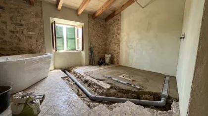 A bathroom mid-renovation shows a plastic-wrapped tub, exposed pipework in a trench, and patterned tiles with cement bags.