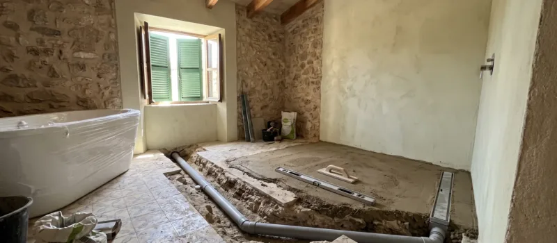 A bathroom mid-renovation shows a plastic-wrapped tub, exposed pipework in a trench, and patterned tiles with cement bags.