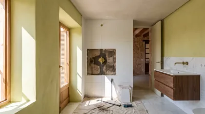 A bathroom under renovation shows a stripped wall patch behind a shower valve, loose tiles, and a crowbar on a dust sheet.