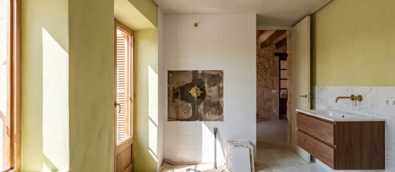 A bathroom under renovation shows a stripped wall patch behind a shower valve, loose tiles, and a crowbar on a dust sheet.