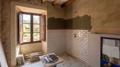 A bathroom mid renovation with subway tiles half laid, red spacer clips, a laser level line and a tiling trowel on a stand.