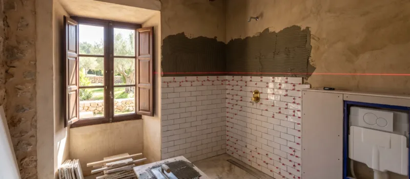 A bathroom mid renovation with subway tiles half laid, red spacer clips, a laser level line and a tiling trowel on a stand.