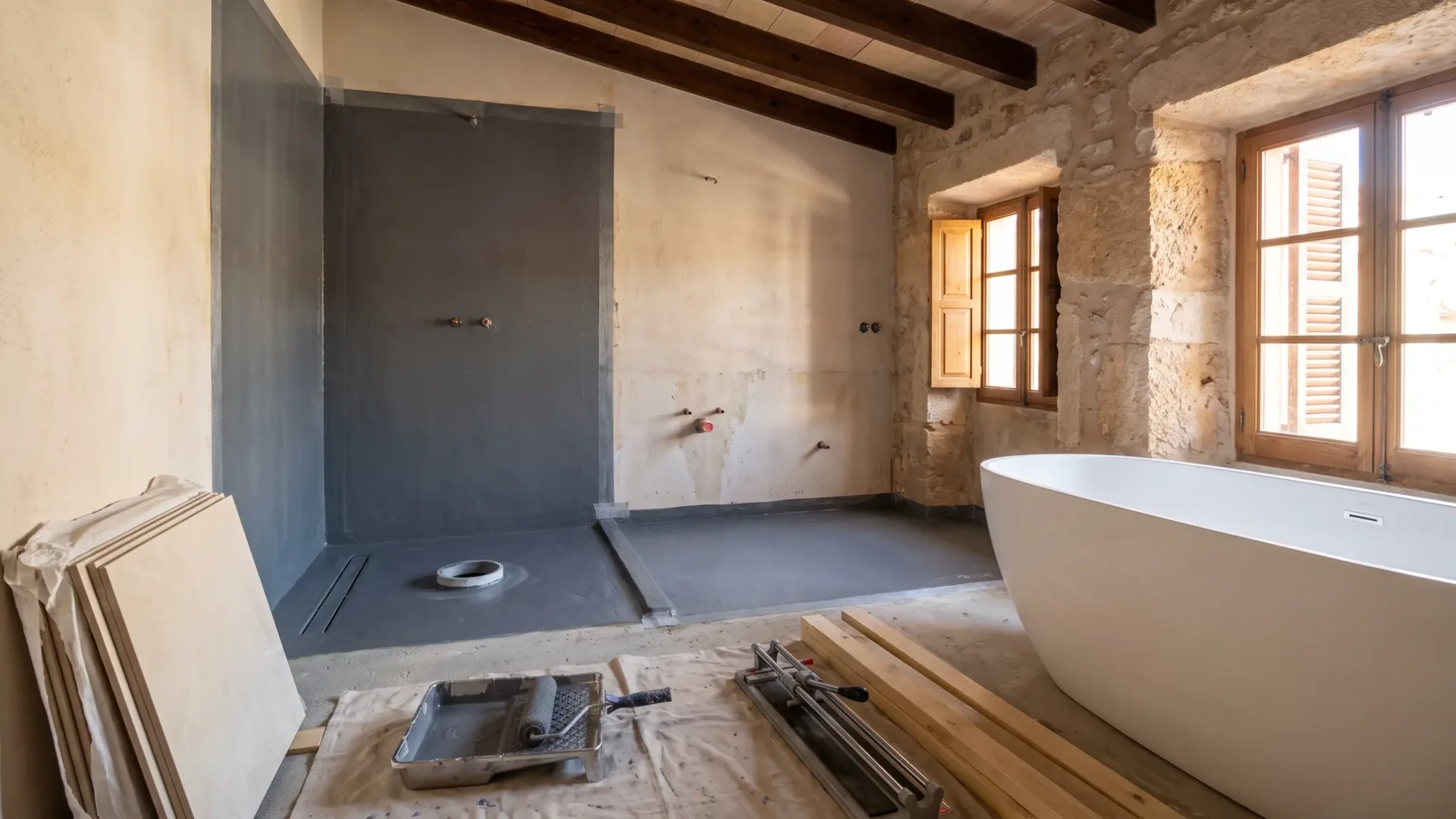 A shower recess mid renovation shows grey waterproofing membrane, exposed pipe stubs, and stacked tiles on the floor.
