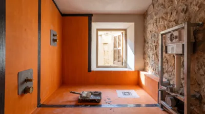 A bathroom mid-fit-out shows orange waterproofing membrane taped across the shower walls, tools left below the window.