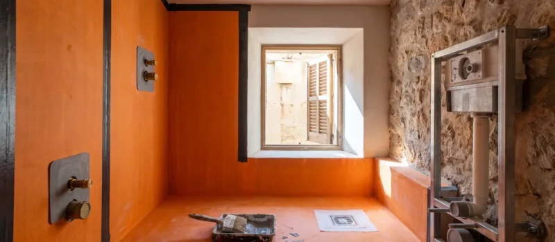 A bathroom mid-fit-out shows orange waterproofing membrane taped across the shower walls, tools left below the window.