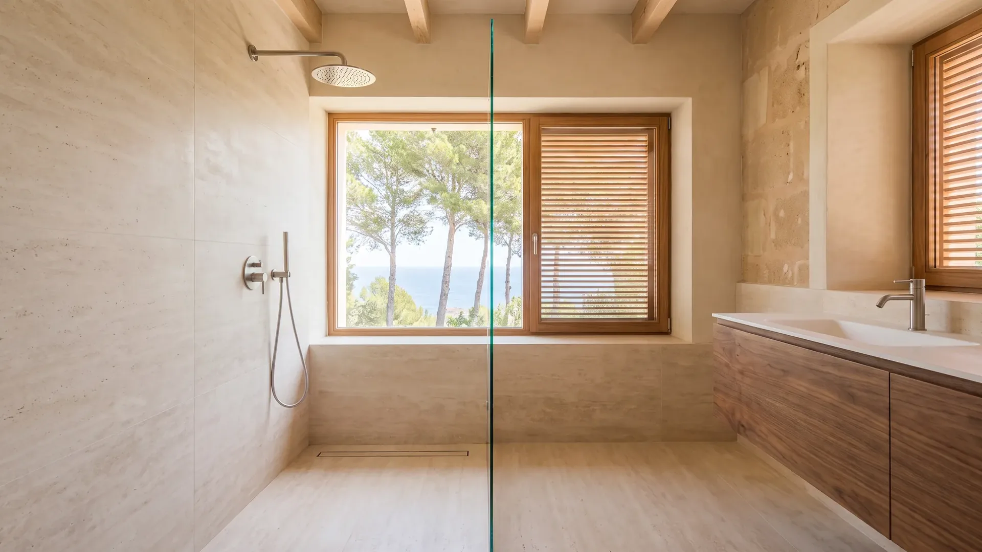 A walk-in shower with travertine tiles faces a window framed by pine trees and the sea, beside a walnut vanity.