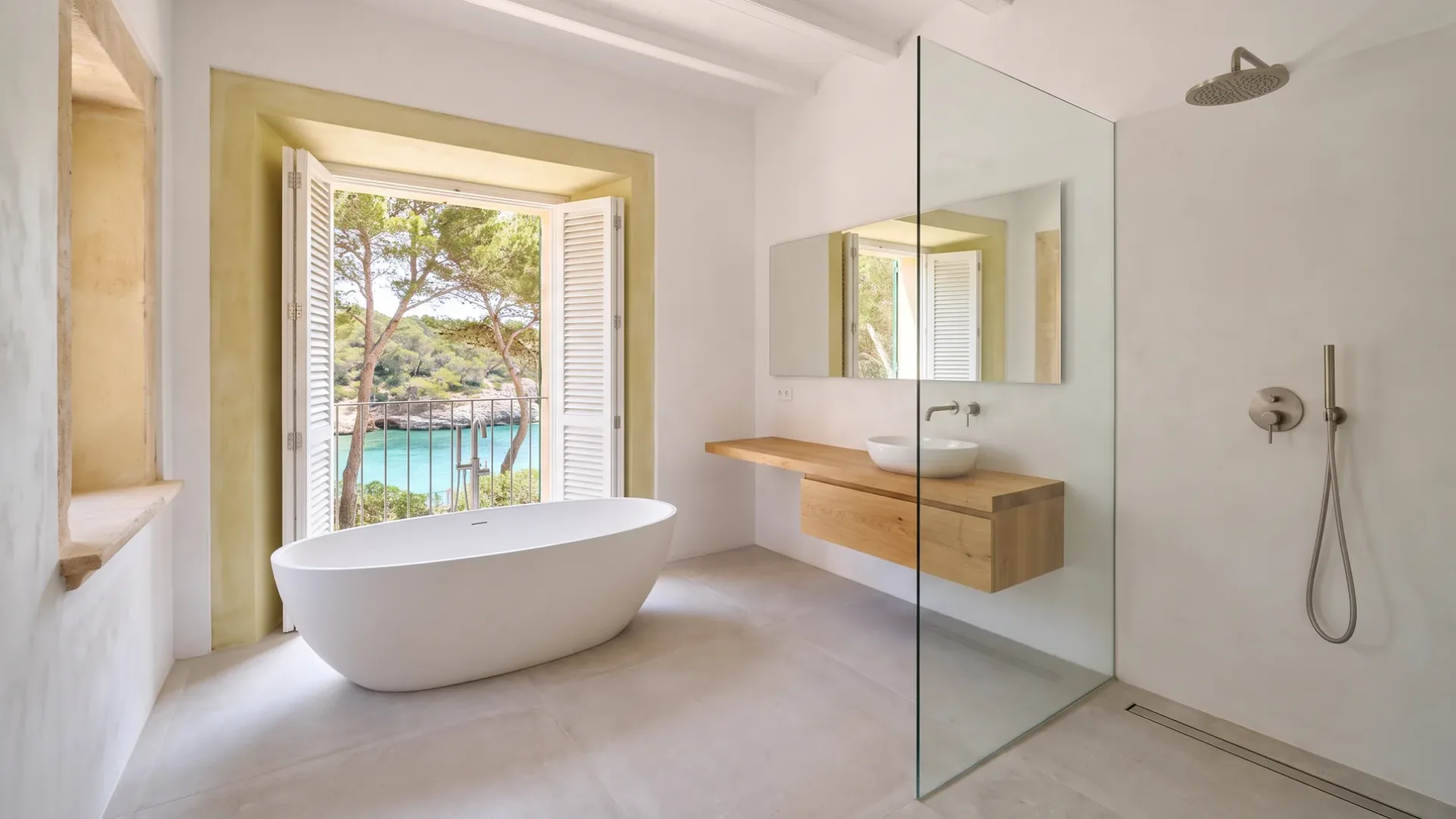 A freestanding oval tub sits before tall white shutters opening onto a sea view, beside a glass shower and wood vanity basin.