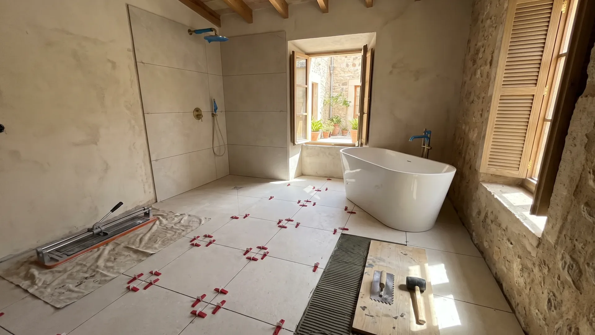 A bathroom mid-renovation shows floor tiles laid with spacers, a trowel and mallet on plywood, and a tub beside the window.