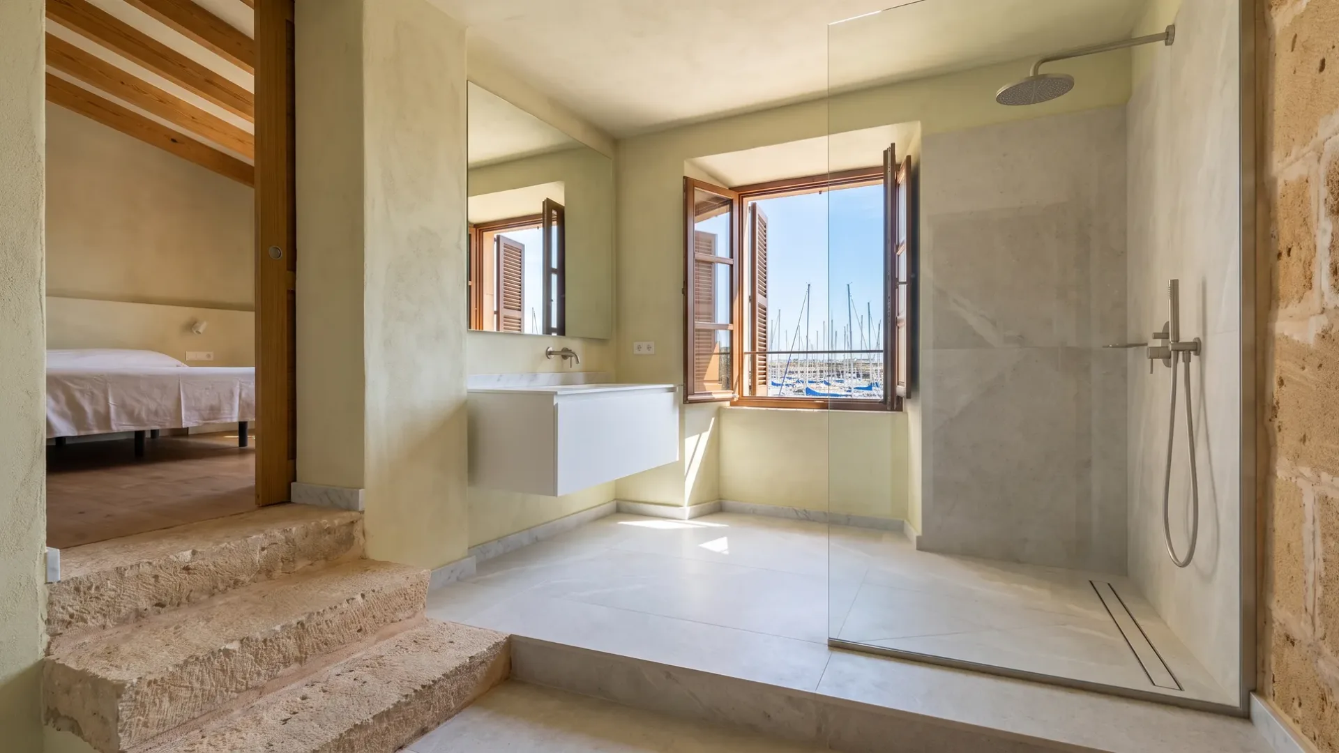 Ein En-Suite-Bad mit einem schwebenden Waschtisch und einer Glasdusche liegt oben auf Steinstufen, das Fenster rahmt einen Jachthafen mit Booten.