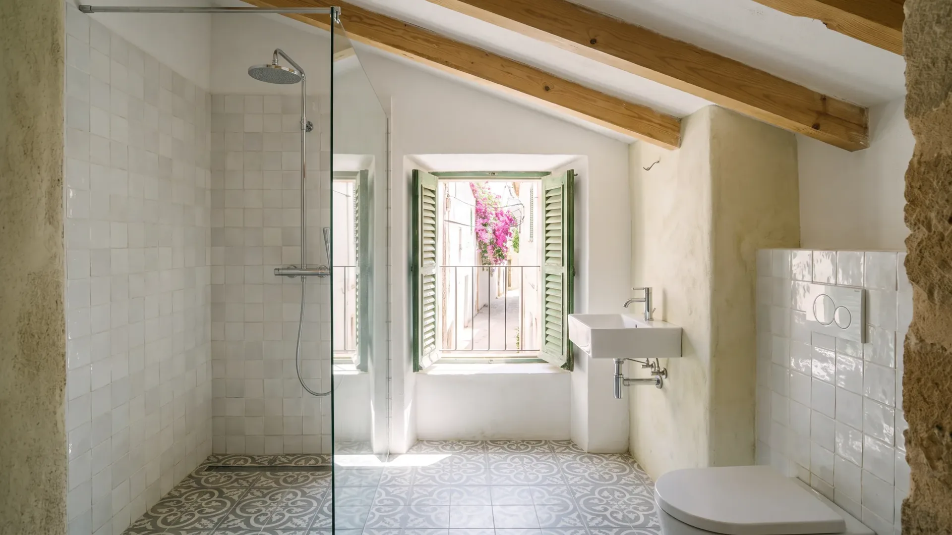 A bathroom with zellige tiles, a patterned floor and a green shuttered window opening on a flowering village street.