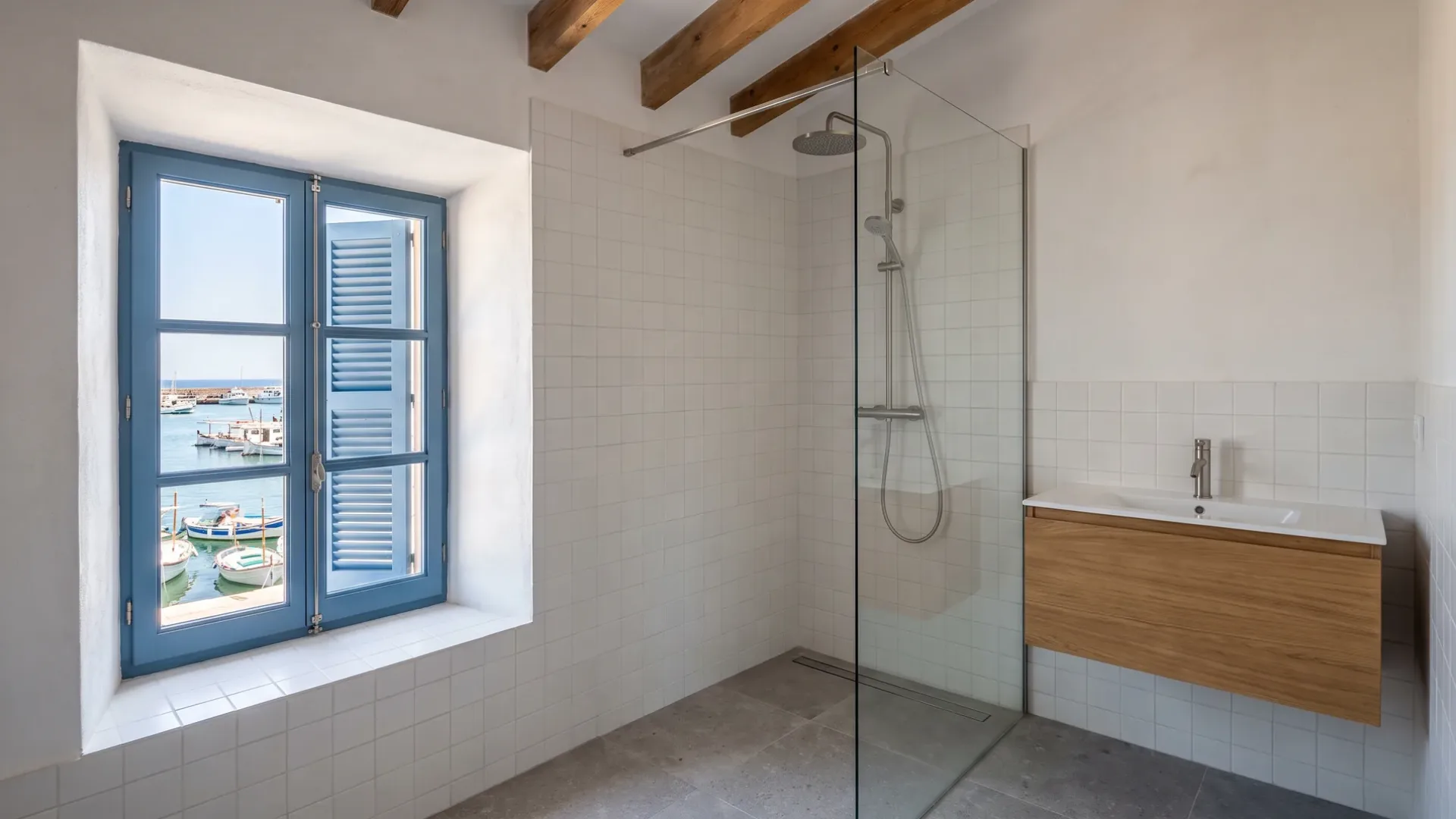 Geflieste Dusche mit Regenbrausekopf und ein Holzwaschtisch stehen neben einem Fenster mit blauen Fensterläden, das Fischerboote im Hafen rahmt.