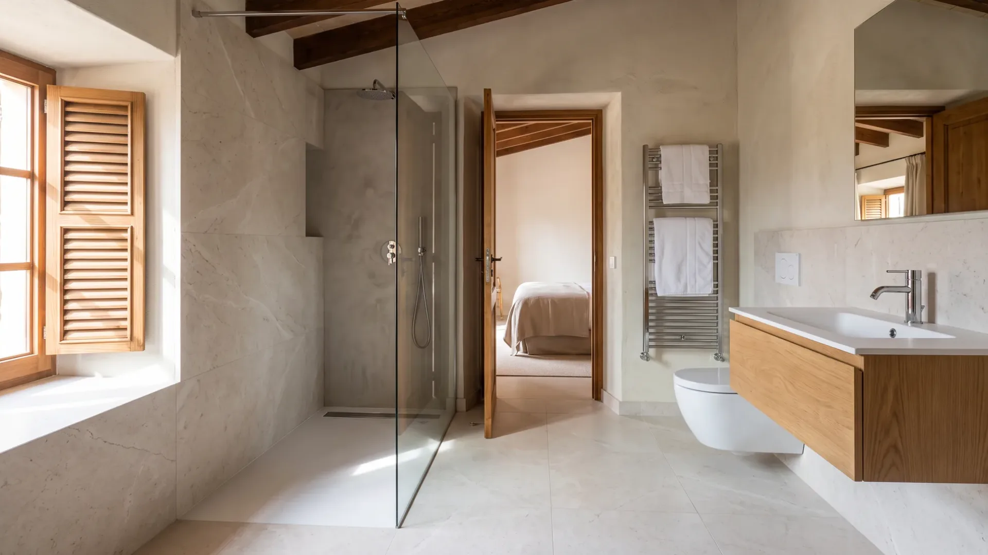A bathroom with a glass walk-in shower, pale stone walls, a chrome towel rail and a wood vanity beside an open bedroom door.