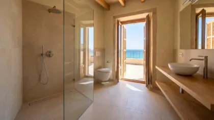 A glass shower with a rain head faces a wall hung toilet and a wooden vanity, beside open shutters framing a sea view.