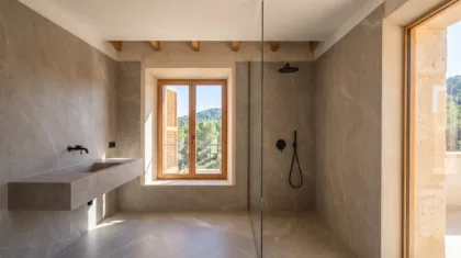 Large format stone effect tiles line a bathroom with a floating basin, a glass shower, and a window over wooded hills.