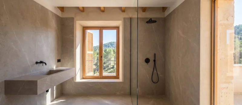 Large format stone effect tiles line a bathroom with a floating basin, a glass shower, and a window over wooded hills.