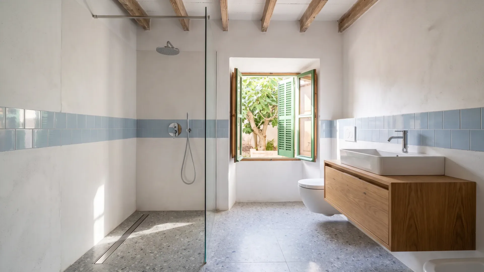 A walk-in shower with a blue tile band and terrazzo floor sits opposite a window framed by a fig tree in a courtyard.