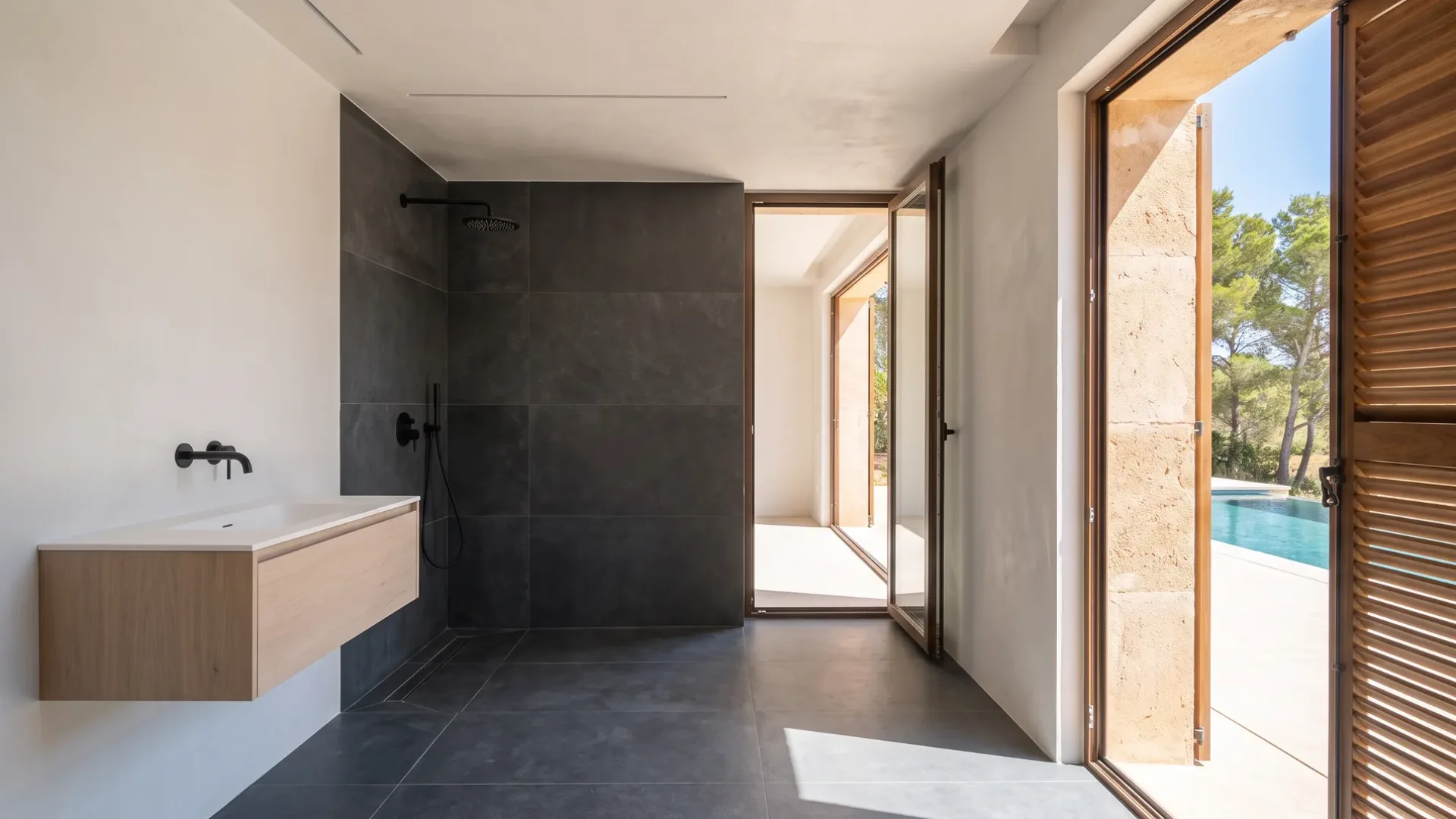 A dark grey tiled shower with black fixtures sits beside a wood vanity, glass doors open to a pool and pine trees outside.