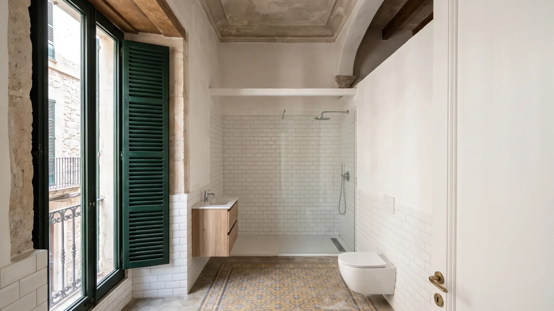 A pale bathroom with a wall hung toilet faces a glass shower lined in white subway tile, under an ornate plaster ceiling.