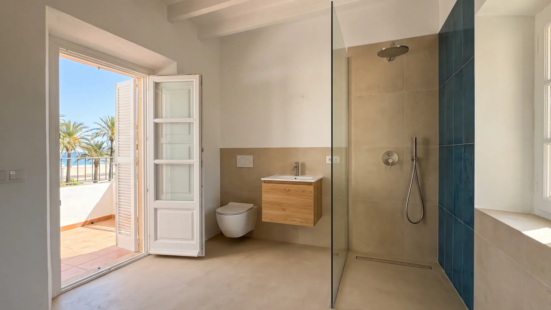 A glass shower with a dark blue tiled accent wall and wood vanity stands beside doors open to a balcony over the sea.