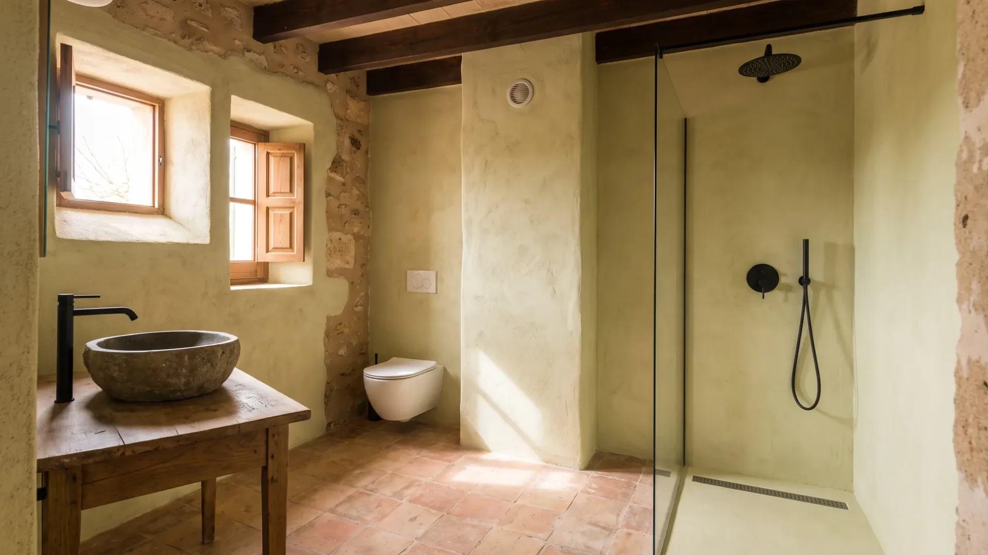 A stone basin on a wooden table sits opposite a toilet and a glass shower with black fittings, under dark ceiling beams.