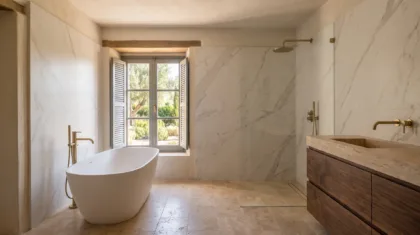 A freestanding oval tub sits before a shuttered window, beside a glass shower and a stone vessel basin on travertine tiles.
