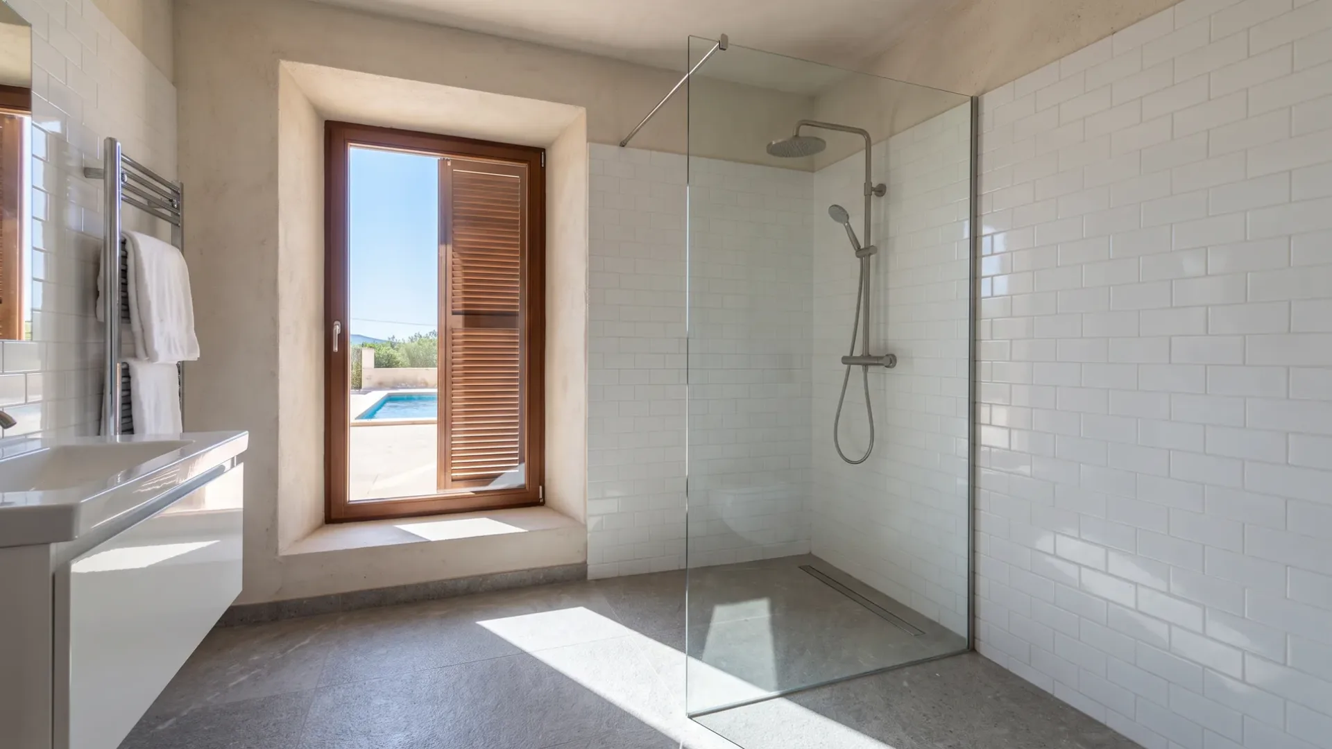 A glass walk in shower lined in white subway tile stands beside a window framing a pool and a wall mounted towel rail.