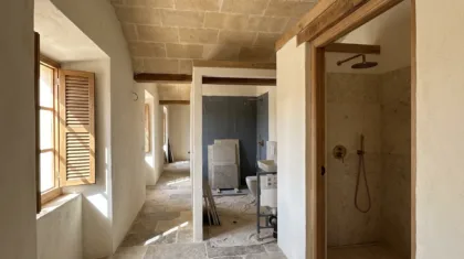 A vaulted stone hallway leads to two bathrooms mid-renovation, one with a tiled wall, another fitted with brass fixtures.