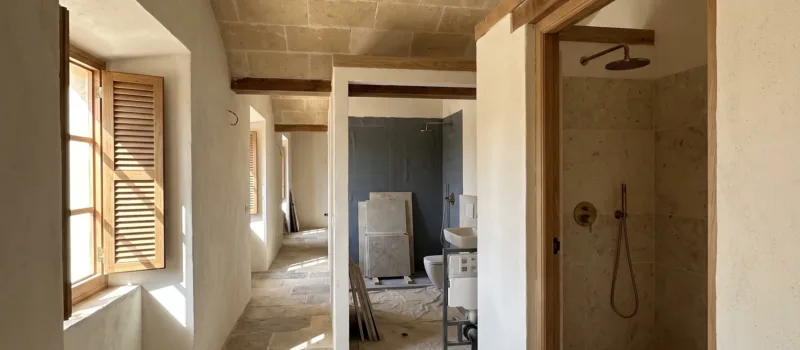 A vaulted stone hallway leads to two bathrooms mid-renovation, one with a tiled wall, another fitted with brass fixtures.
