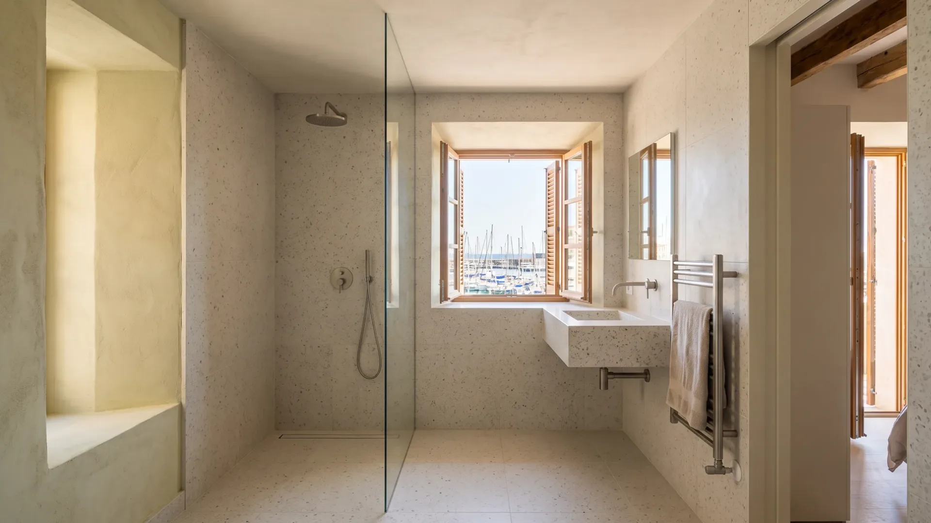 A speckled stone-tiled shower and floating basin sit beneath a timber window, overlooking a marina full of boats.