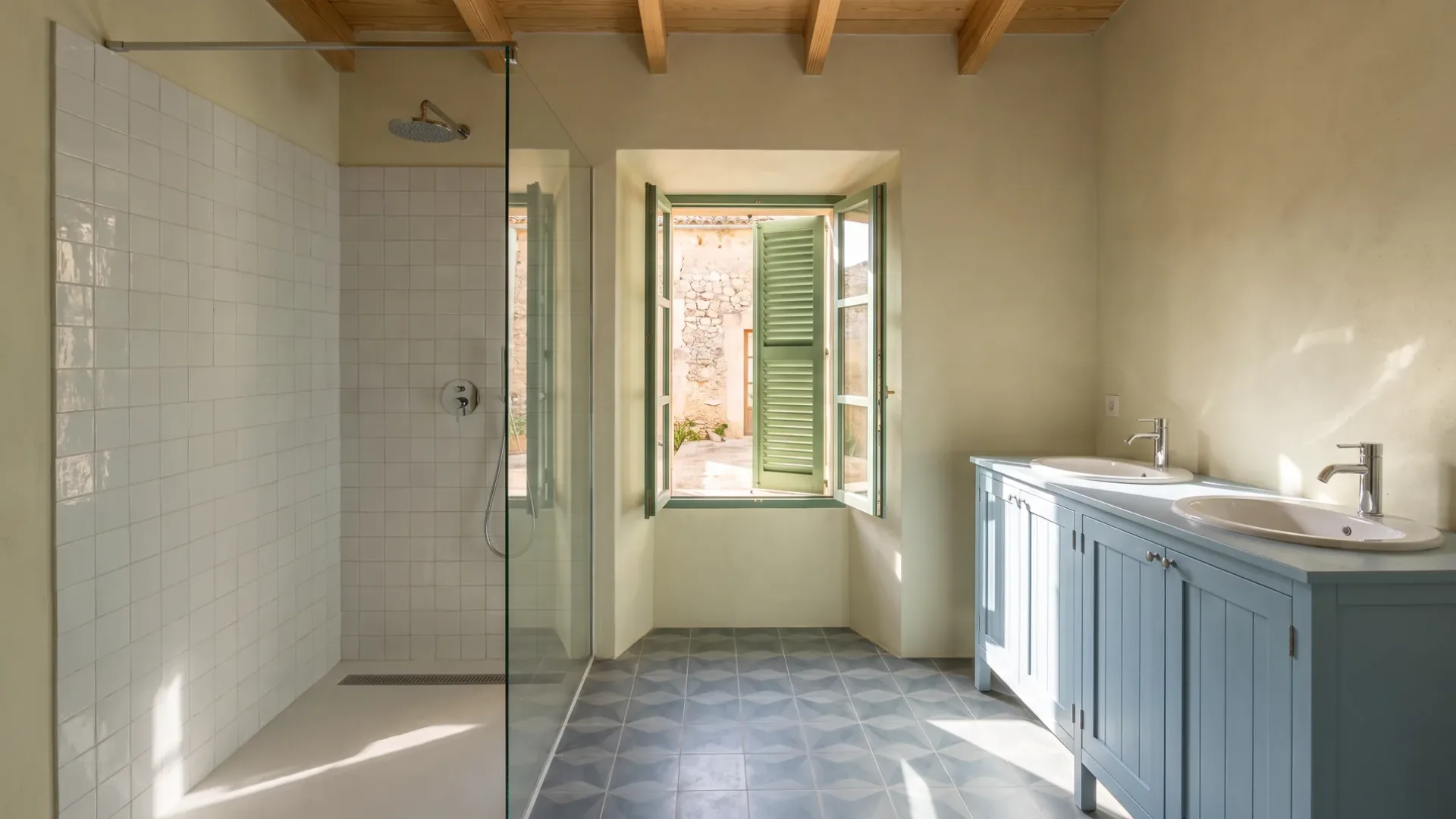 A bathroom with a white tiled shower, patterned floor tiles and a pale blue double vanity beside a green shuttered window.