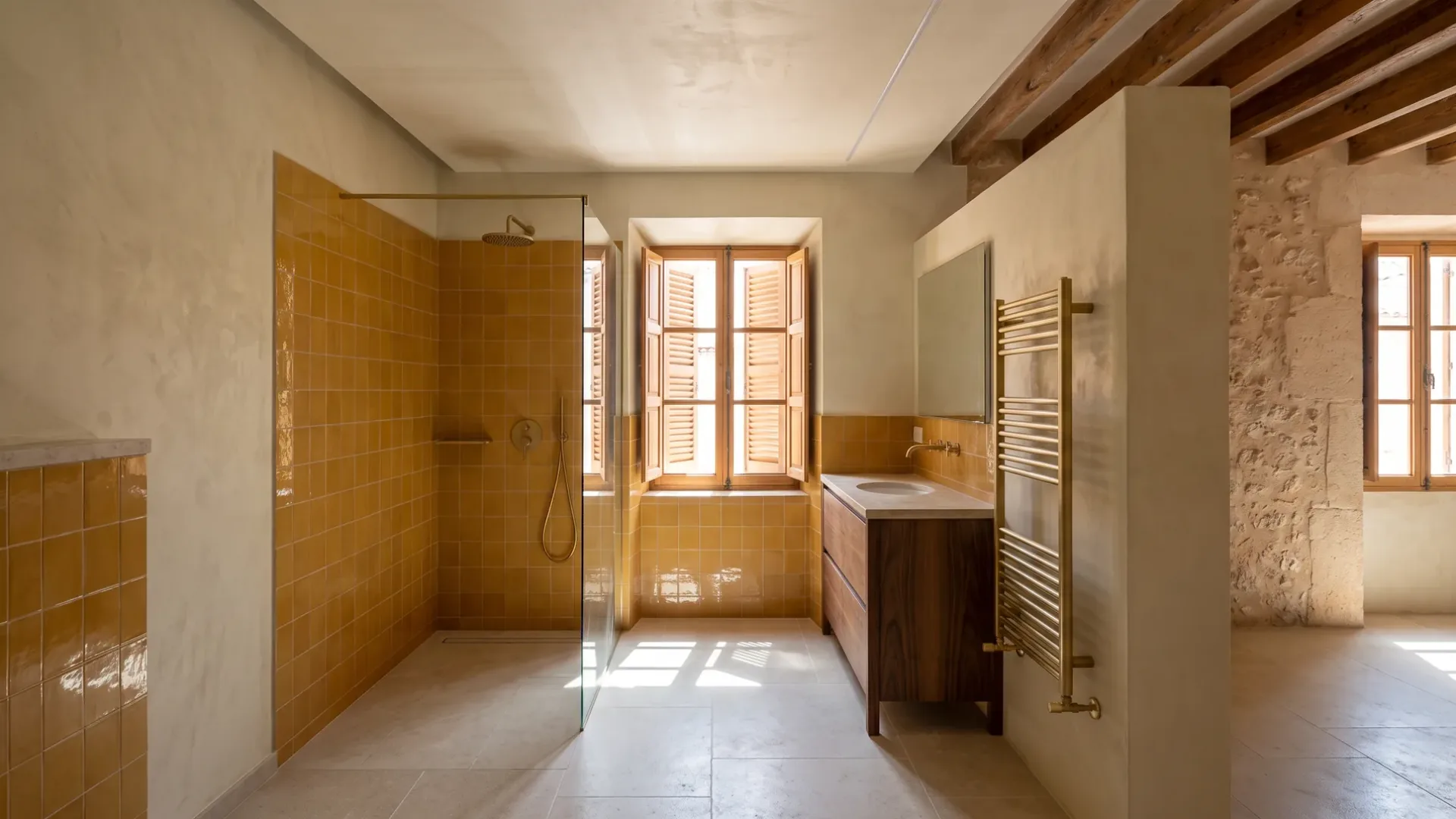A mustard tiled walk-in shower sits beside a wood vanity and a brass towel rail, with a stone wall seen through a doorway.