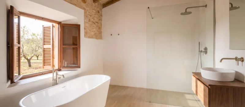 A freestanding tub with floor mounted taps sits beneath a window opening onto orchard trees, opposite a glass walk in shower.