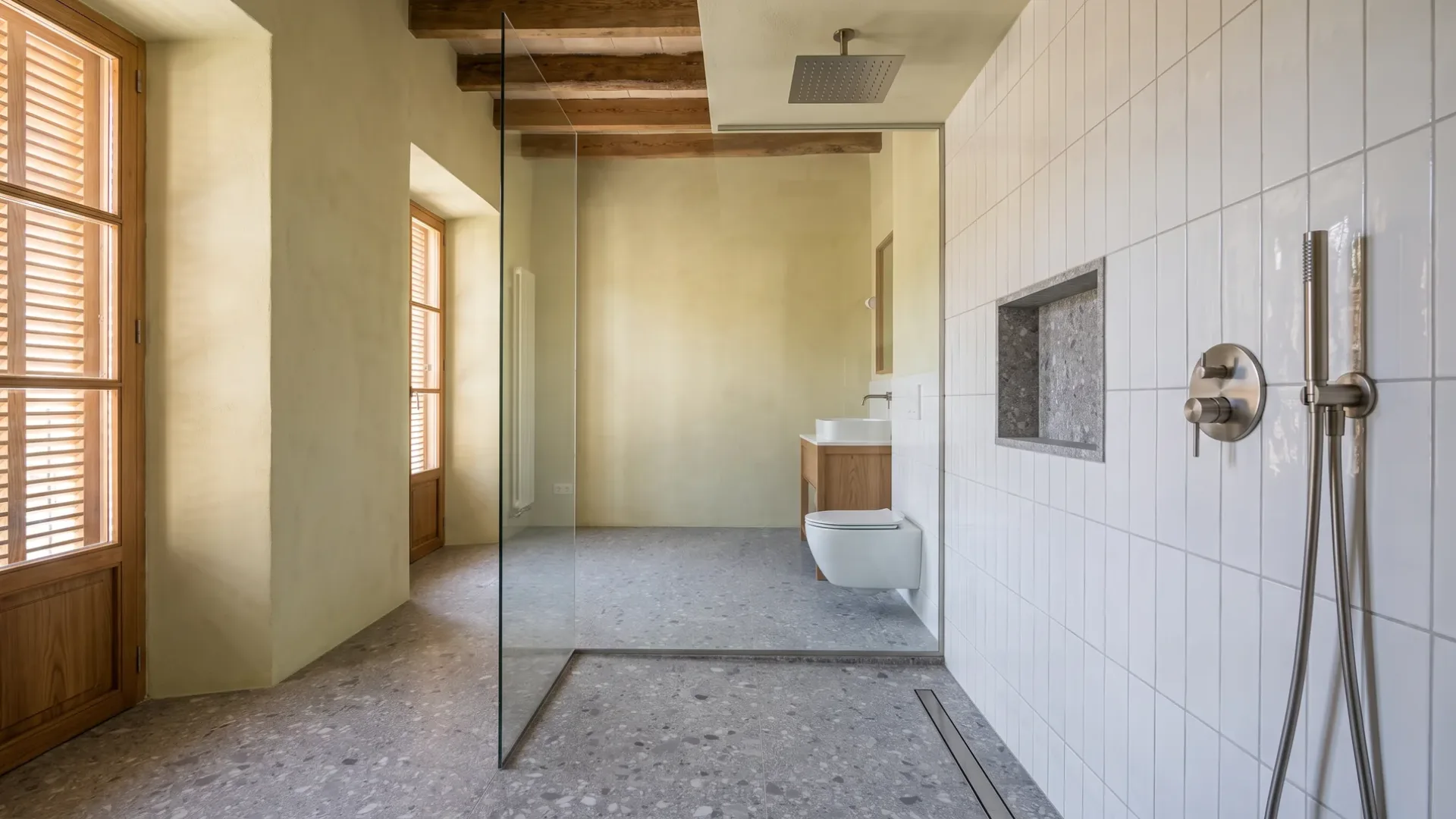 A glass-walled walk-in shower with terrazzo floor faces a white tiled wall with a stone niche, near a wood vanity and toilet.