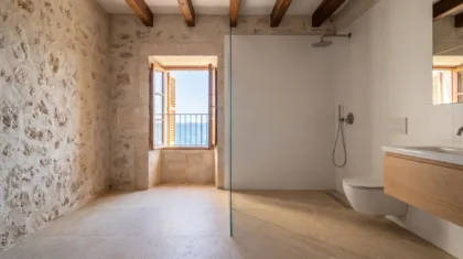 A stone walled wet room with white tiles and a wood vanity opens to a window with a sea view and balcony railing.
