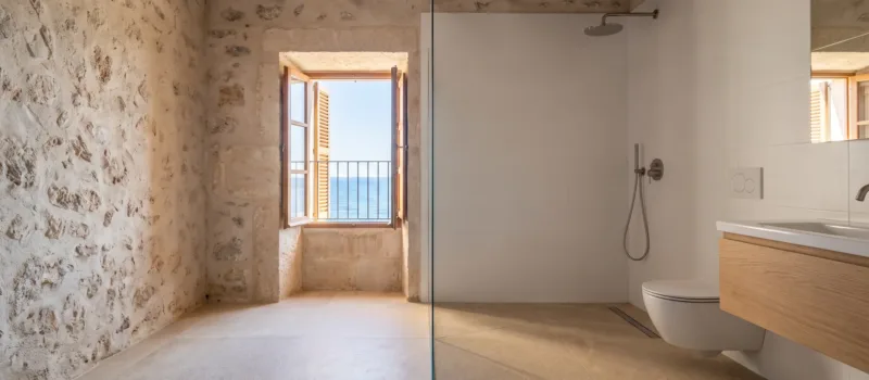 A stone walled wet room with white tiles and a wood vanity opens to a window with a sea view and balcony railing.