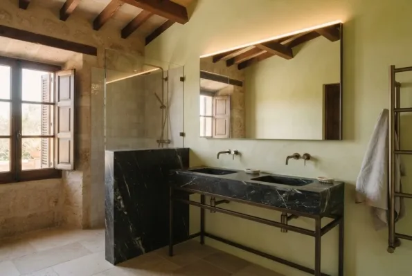 A bathroom with a black marble double basin on a bronze frame, a backlit mirror, and a glass shower against a stone wall.