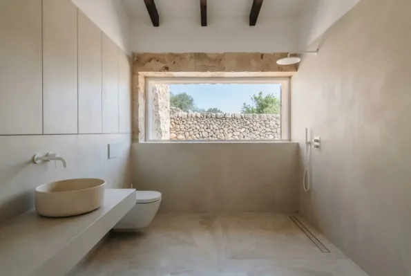 A pared-back bathroom with flush plaster cabinets, a terrazzo basin and a long window framing a dry stone wall outside.
