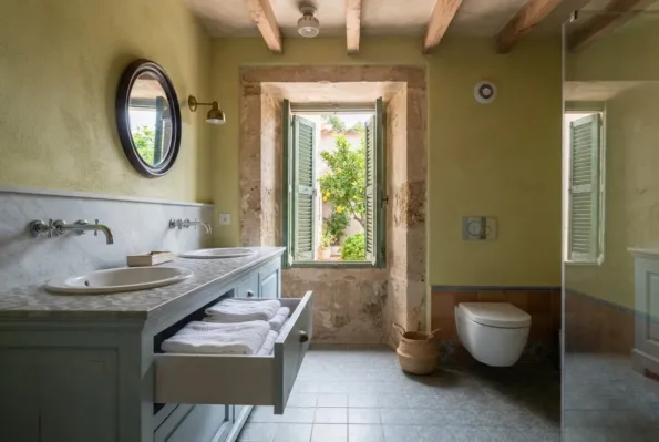 A bathroom with a marble topped double vanity, an oval mirror, a wall hung WC, and shutters open to a lemon tree.