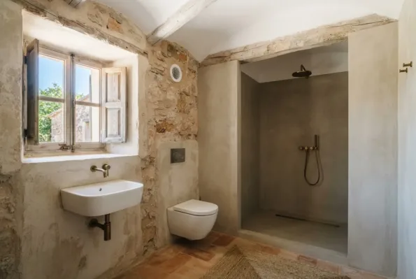 Ein Steinbad hat ein rundes Wandlüftungsgitter über einem Waschbecken, ein WC und eine verputzte Duschnische mit Messingarmaturen.