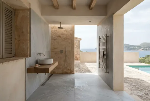 Un baño de obra abierto a una terraza con piscina y vistas al mar, con una ducha de paredes de piedra, un lavabo sobre balda de madera y un toallero.